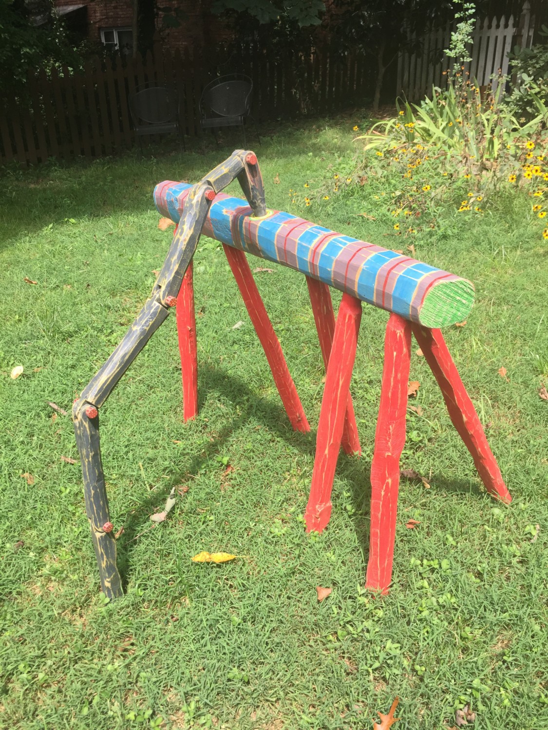 Sawhorse-like sculpture with a striped blue and red log on tall red legs, with a bent black articulated arm