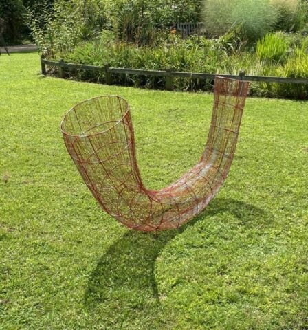 Curved sculpture made of red wire mesh, displayed outdoors on grass