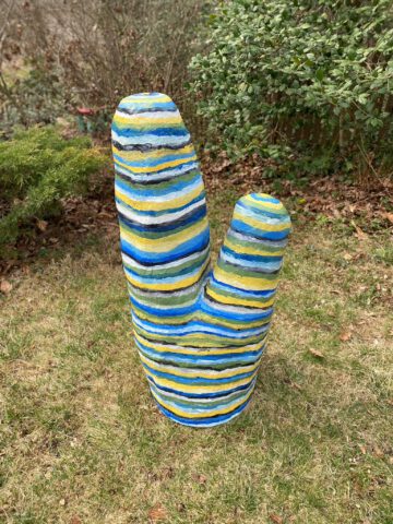 Two tall narrow sculptures with horizontal blue, yellow, green, and white stripes, standing in grass among garden shrubs.