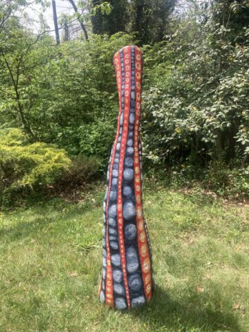 Tall tapering sculpture with red and gray spotted stripes, standing in grass